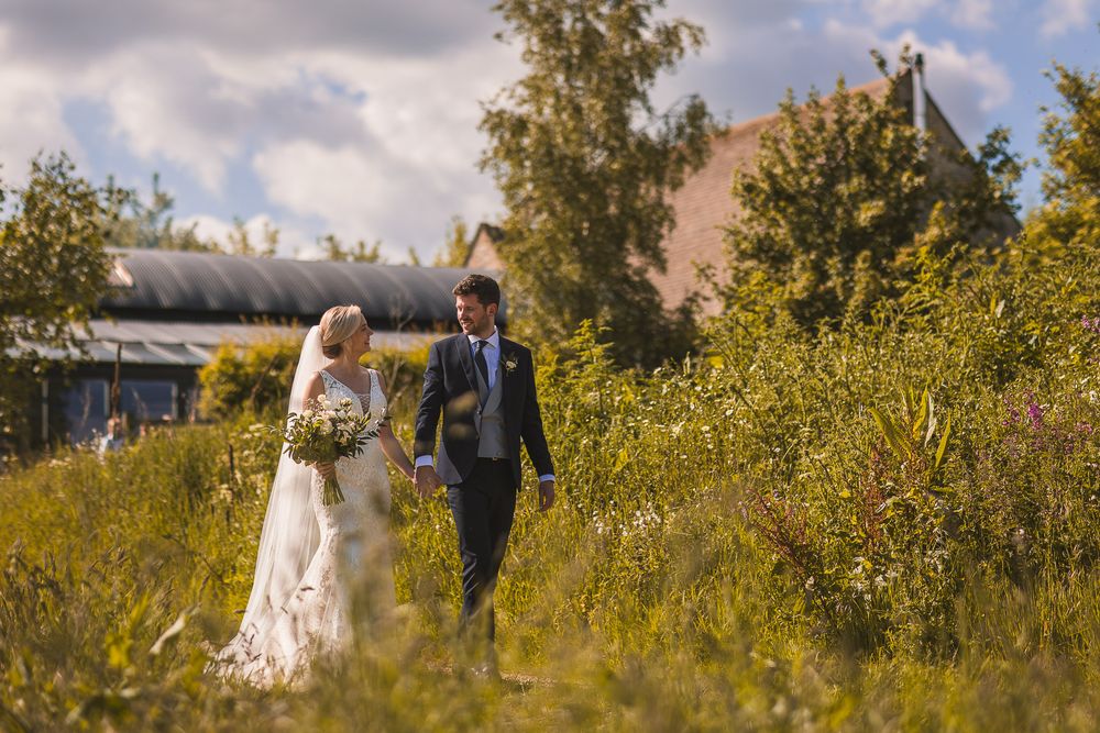 A bridal couple walks hand in hand through a scenic field of yellow wildflowers on a sunny autumn day at Stone Barn in the Cotswolds.