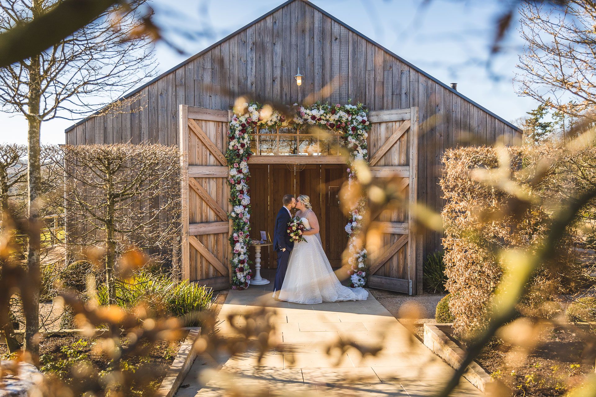 A bride and groom pose outside the rustic wooden barn at Hyde House with open doors glowing in warm sunset light against a blue sky.