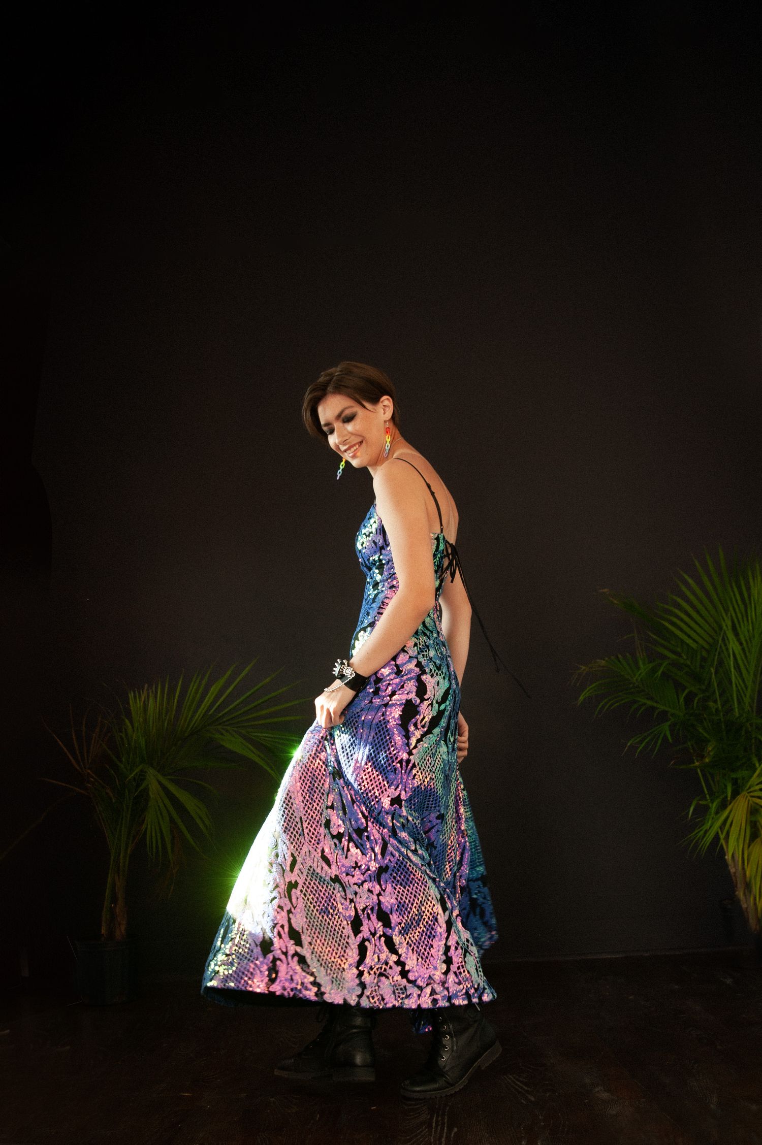 A fashion model twirls in a shimmering blue and purple evening gown against a dark background with palm plants.