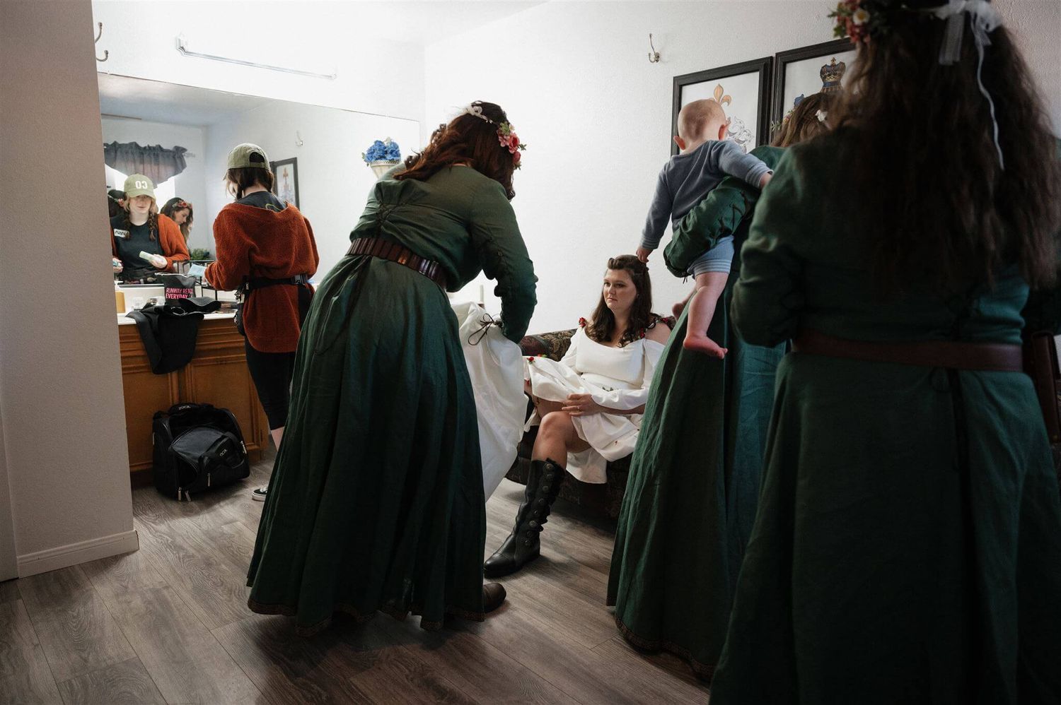 People in medieval-style green dresses gather in a preparation room with white walls and wood flooring.