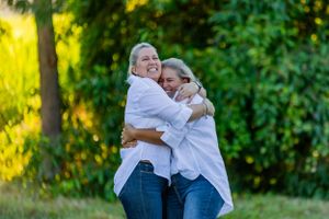 Two people share a warm embrace while laughing outdoors in matching white shirts.