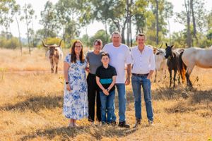 A small family group poses for a photo with livestock and eucalyptus trees in the rural background.