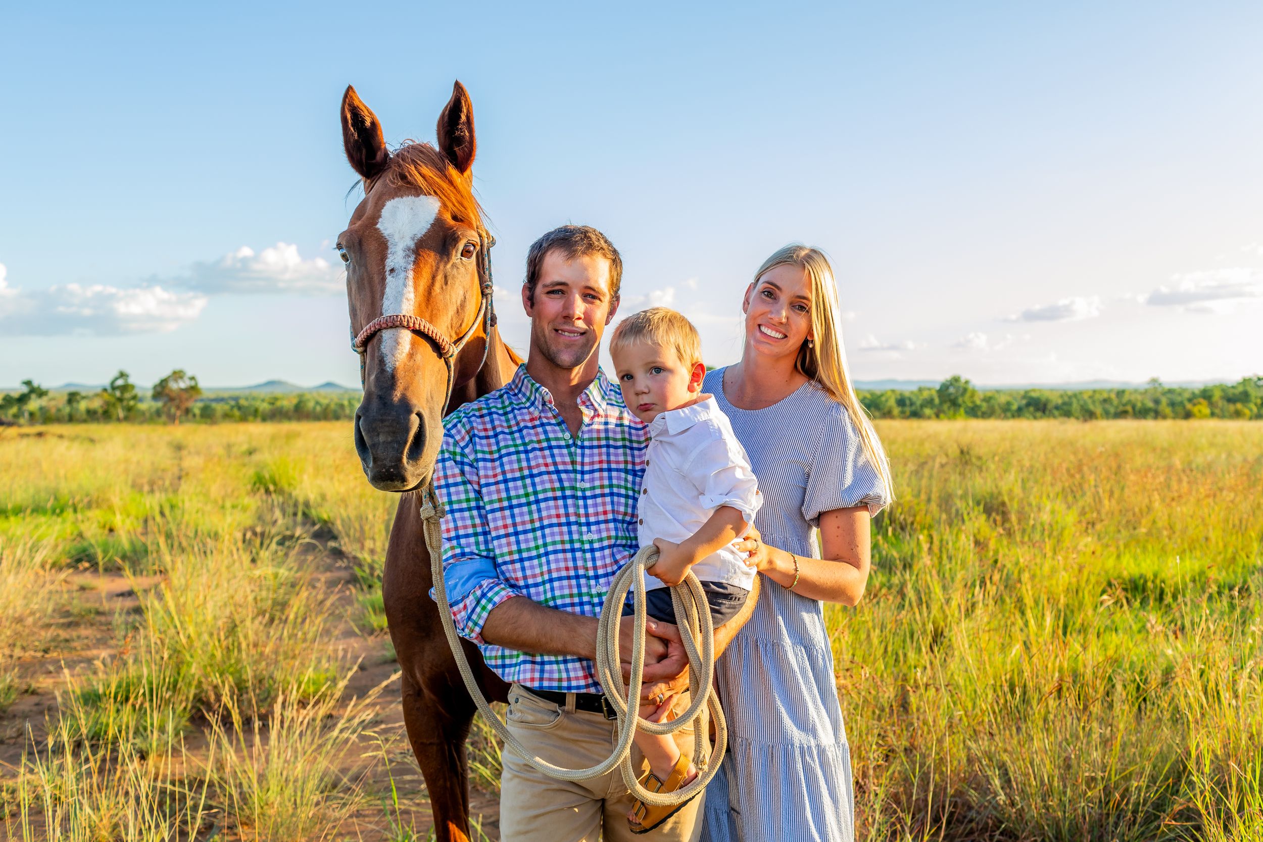 Family standing together with brown horse in sunlit meadow.