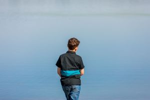 A person in black clothing stands alone looking out over calm blue water.