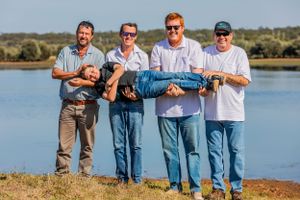 Four people playfully hold another person horizontally by a waterfront on a sunny day.