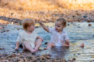 Two small children sit and play together in shallow water at the edge of a stream.