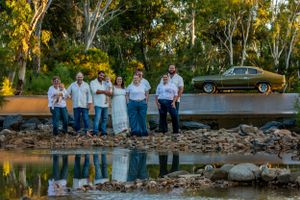 A group in coordinating blue and white outfits poses near a classic car by a reflective pond.