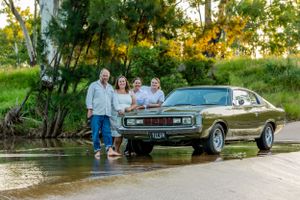 A classic brown muscle car parked by a pond with people standing beside it.