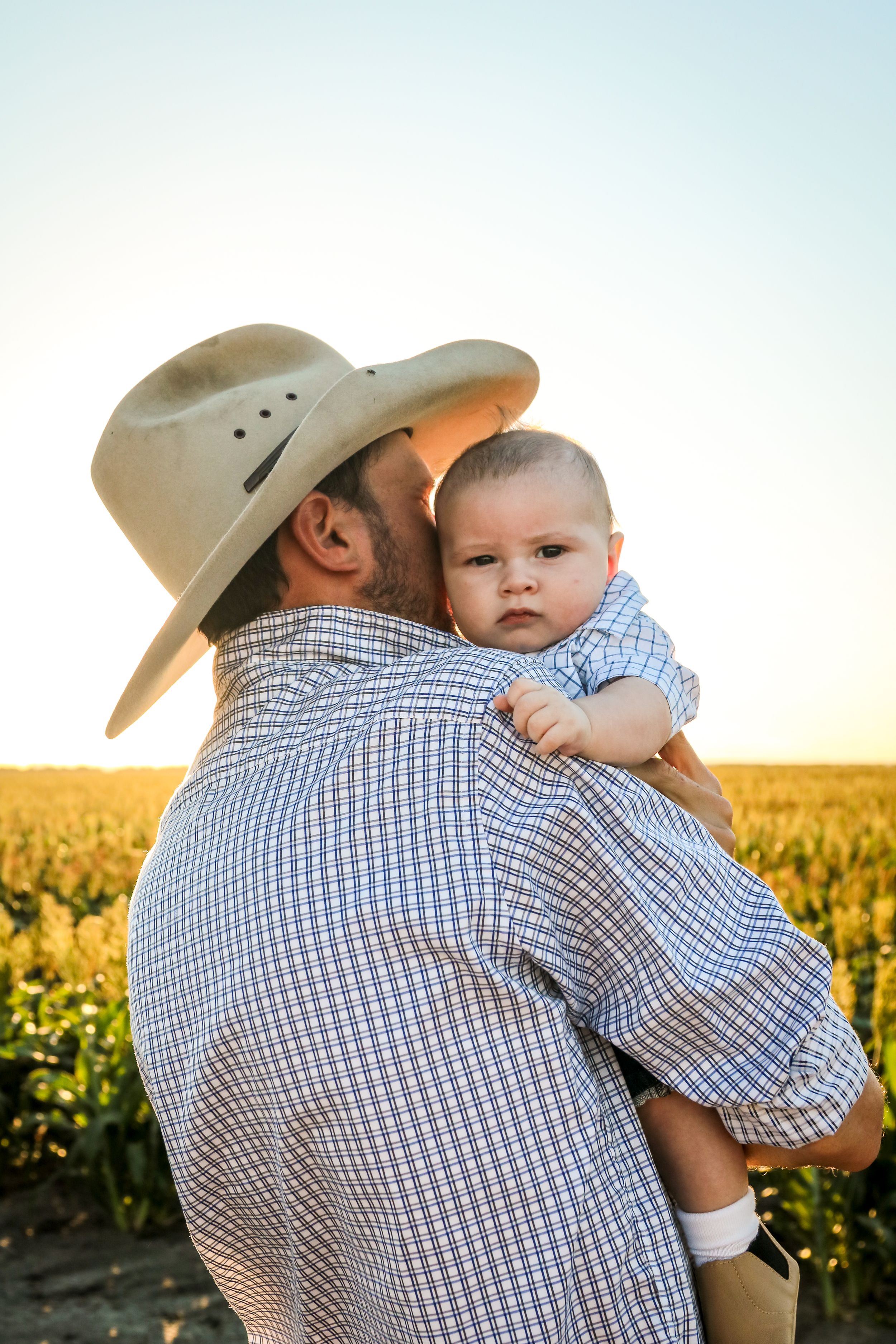 A person wearing a cowboy hat holds a baby in a plaid shirt against a backdrop of a sunny agricultural field.