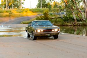A vintage brown muscle car driving on a wet road near water during golden hour.