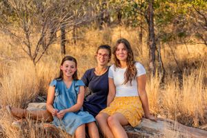 Three people sit together on a rock in a golden-lit natural setting with dry grass and trees.