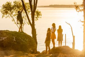 Three silhouettes stand against a golden sunset by a lake with trees framing the peaceful scene.