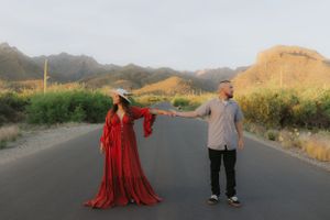 Couple in contrasting outfits posing on desert road with mountains in background.