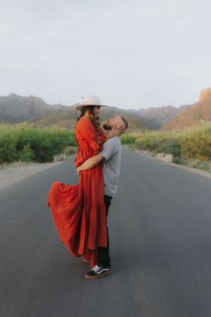 Romantic lift pose on desert road with mountains and sunset in background.