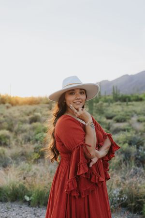A person in a flowing red dress and wide-brimmed hat poses in a desert landscape with mountains and cacti at sunset.