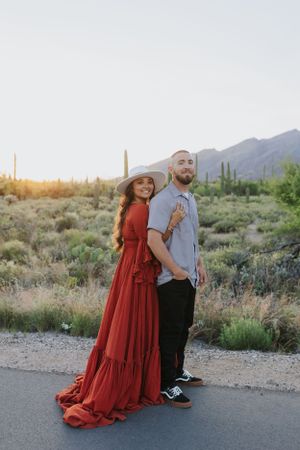 A couple poses playfully on a desert road with mountains and saguaro cacti in the background.