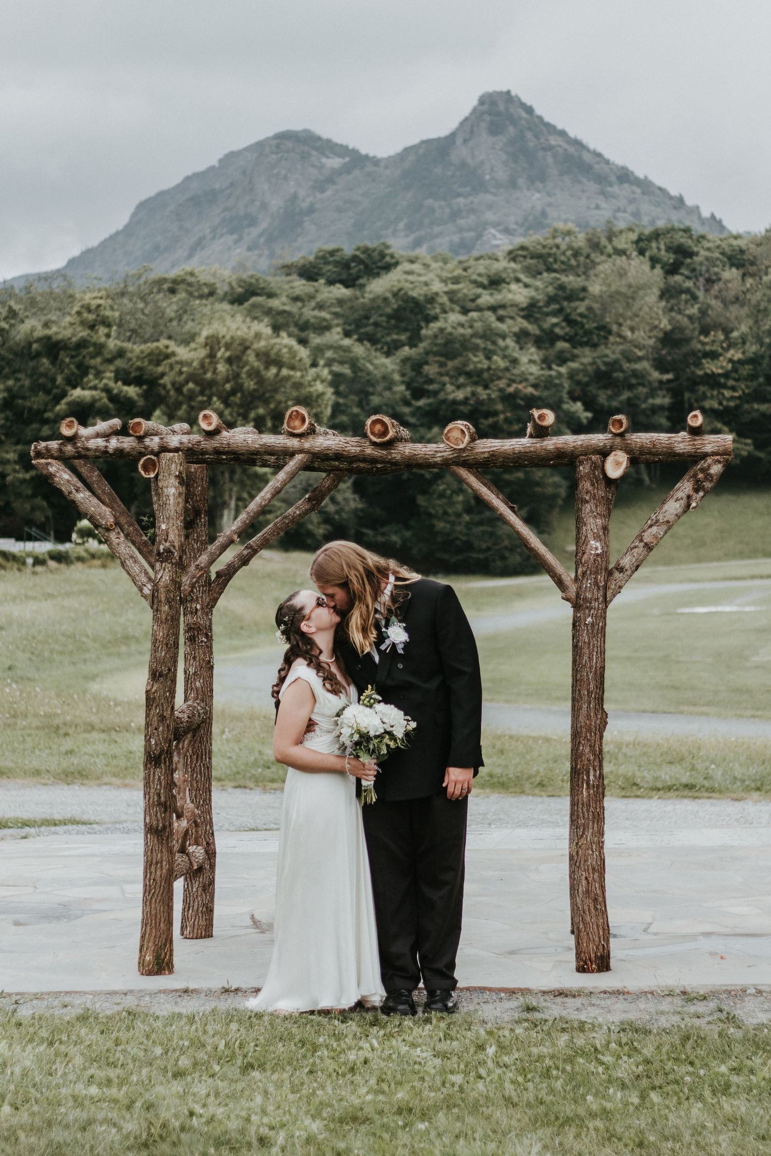 Wedding ceremony under rustic wooden arch with dramatic mountain backdrop.
