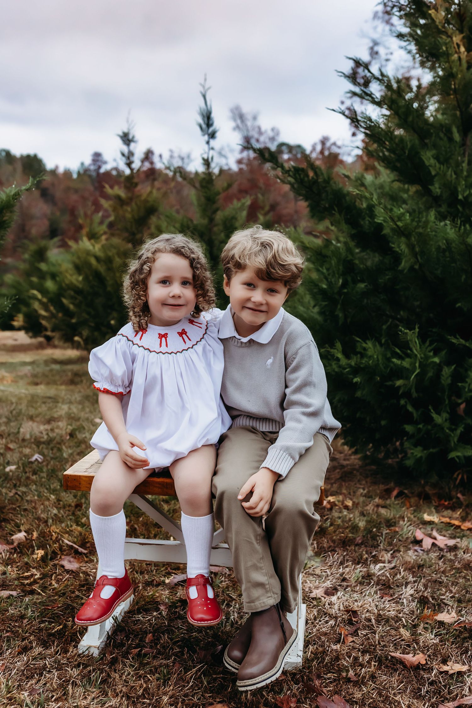 Two children sitting together on a wooden bench at a Christmas tree farm in seasonal holiday outfits.
