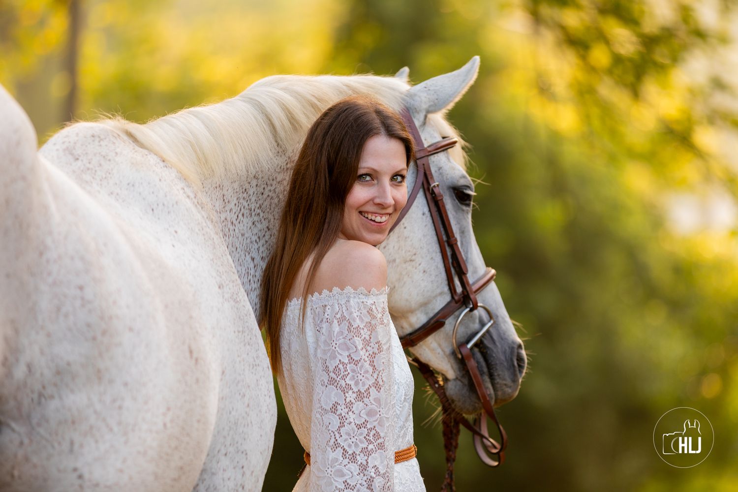 A beautiful white horse with bridle poses with someone in a lace dress during a sunset photo shoot.