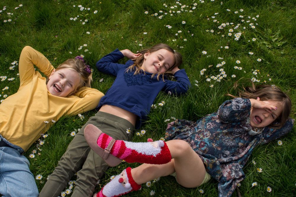 Three children sprawled on lush green grass, heads together, giggles filling the air at Jesus Green park