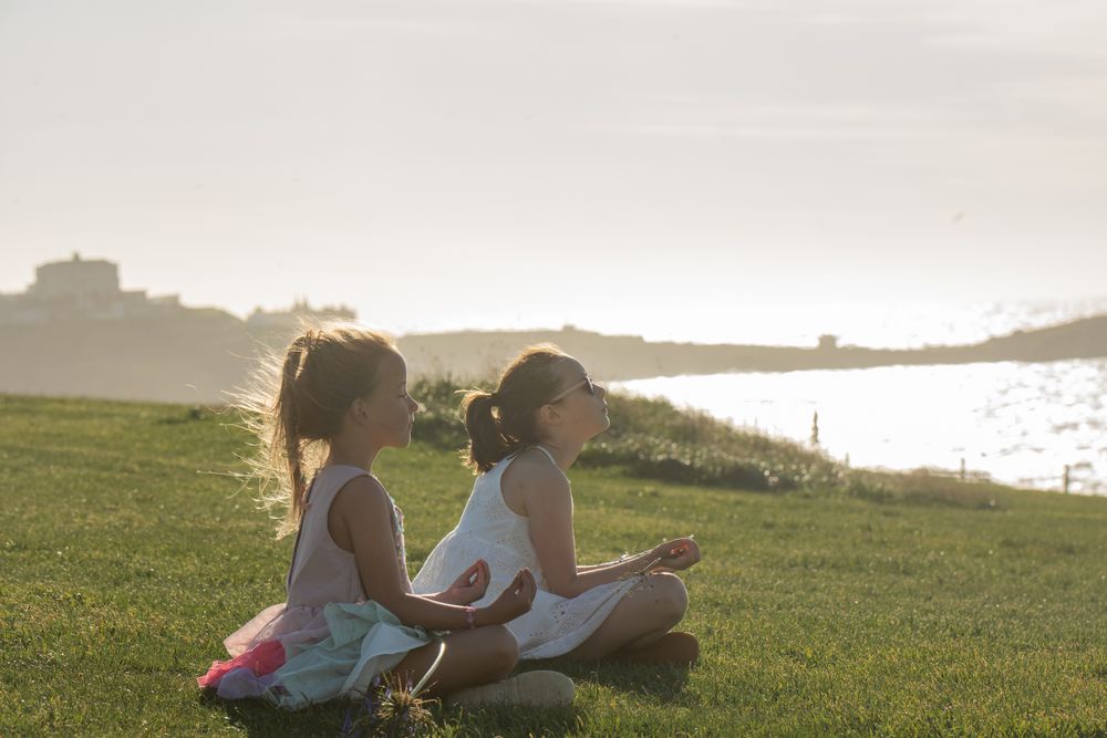 Two children sitting in lotus position meditating on a grassy hill overlooking a sunlit lake at dusk in Cornwall, UK