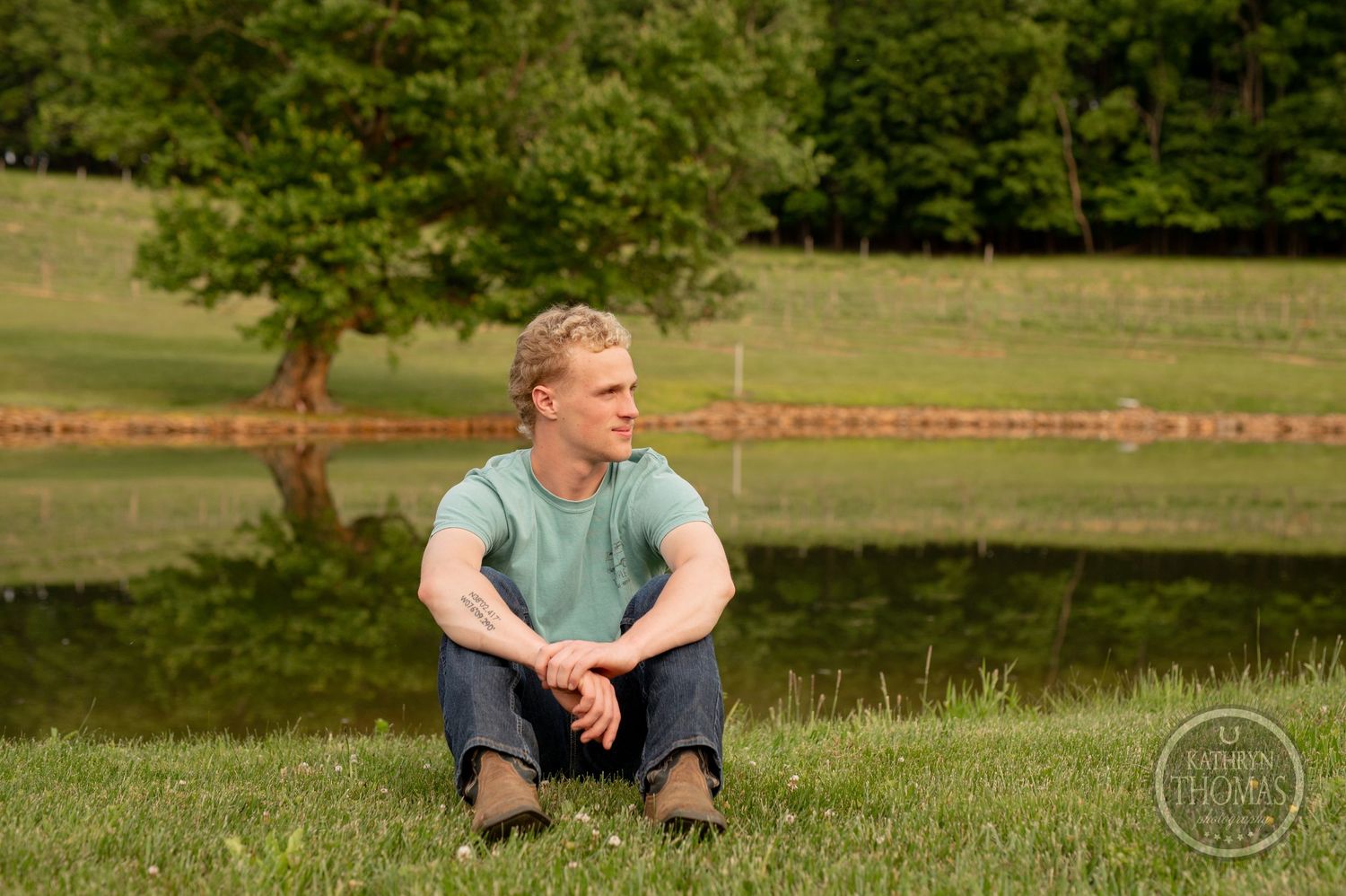 Person in mint shirt sits casually on hillside with pond view.