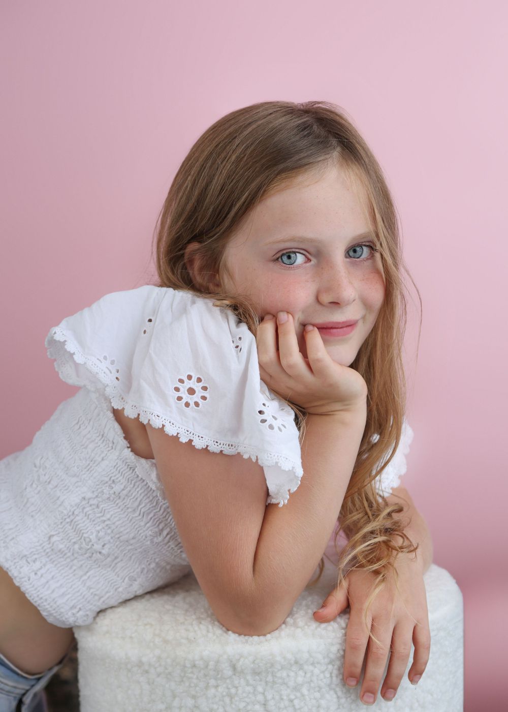 Young person in white eyelet dress poses against pink background with dreamy expression.