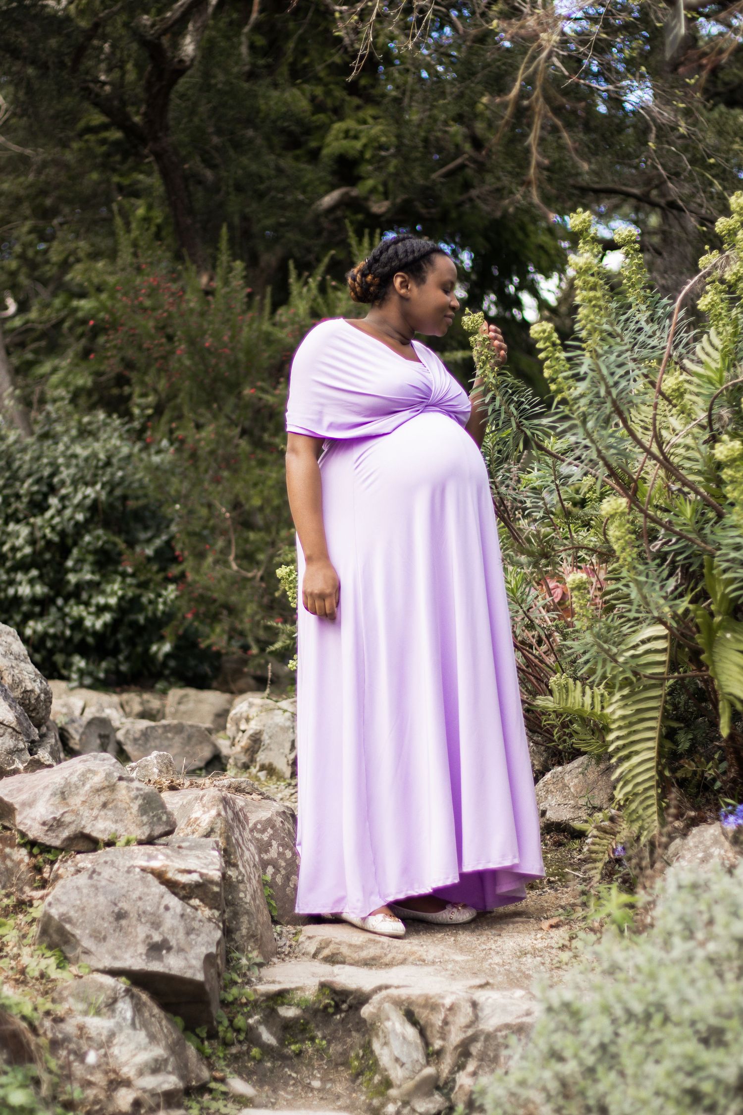 Maternity photo of person in flowing lilac dress standing on stone steps in garden setting.