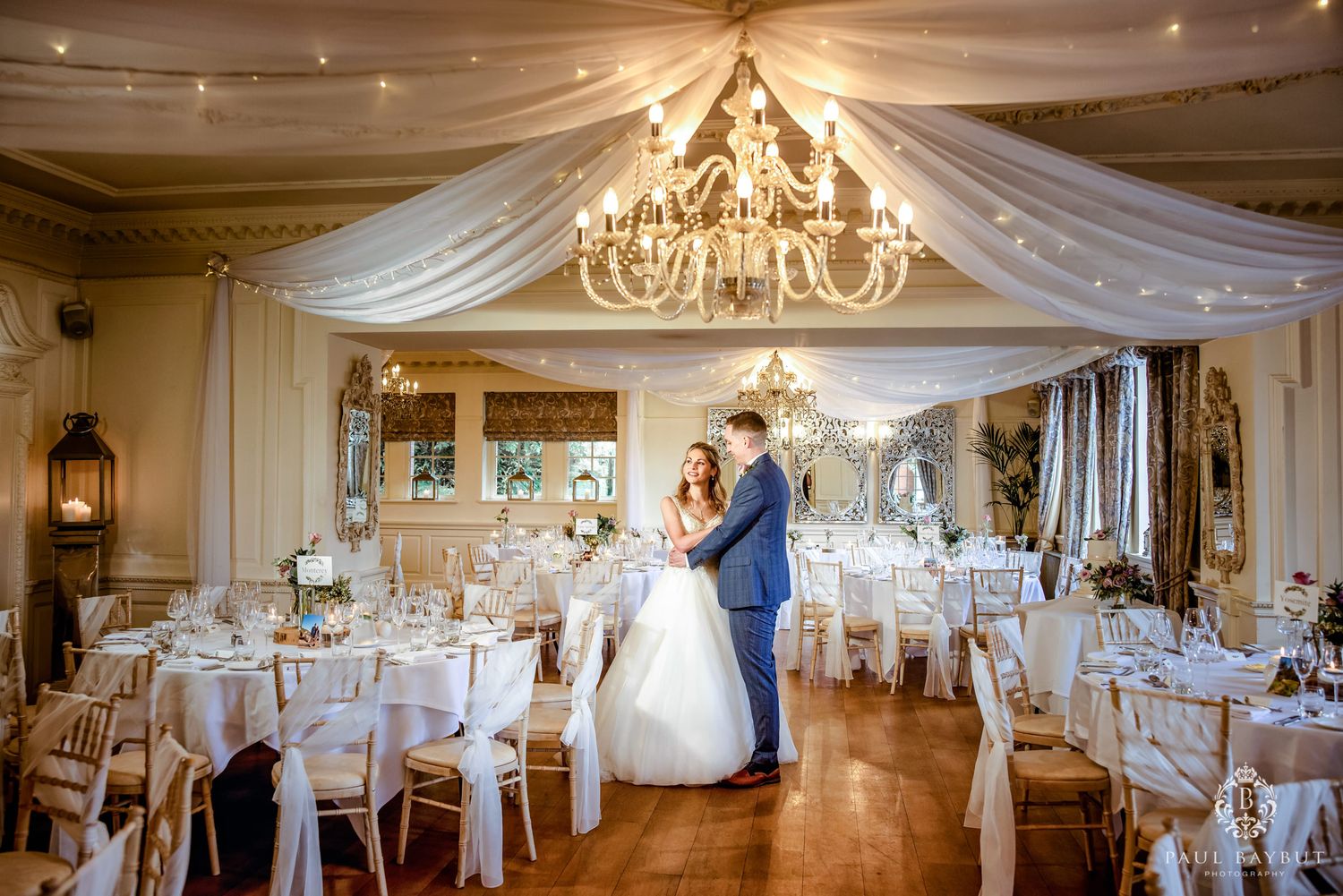 Elegant ballroom with draped ceiling, crystal chandelier and white table settings for a formal wedding reception.