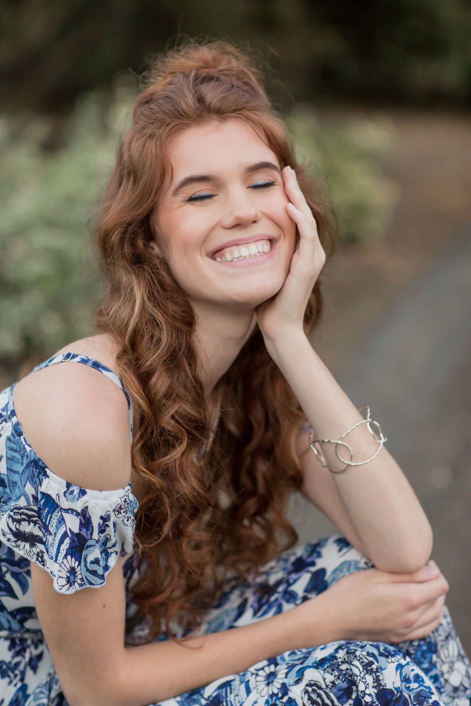 Laughing redhead young woman in blue floral dress with silver bracelet touching her face.