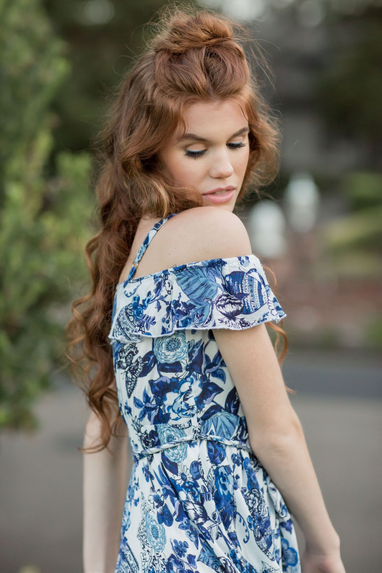 Young redhead woman with half-up curly hair in blue floral off-shoulder dress looking down.
