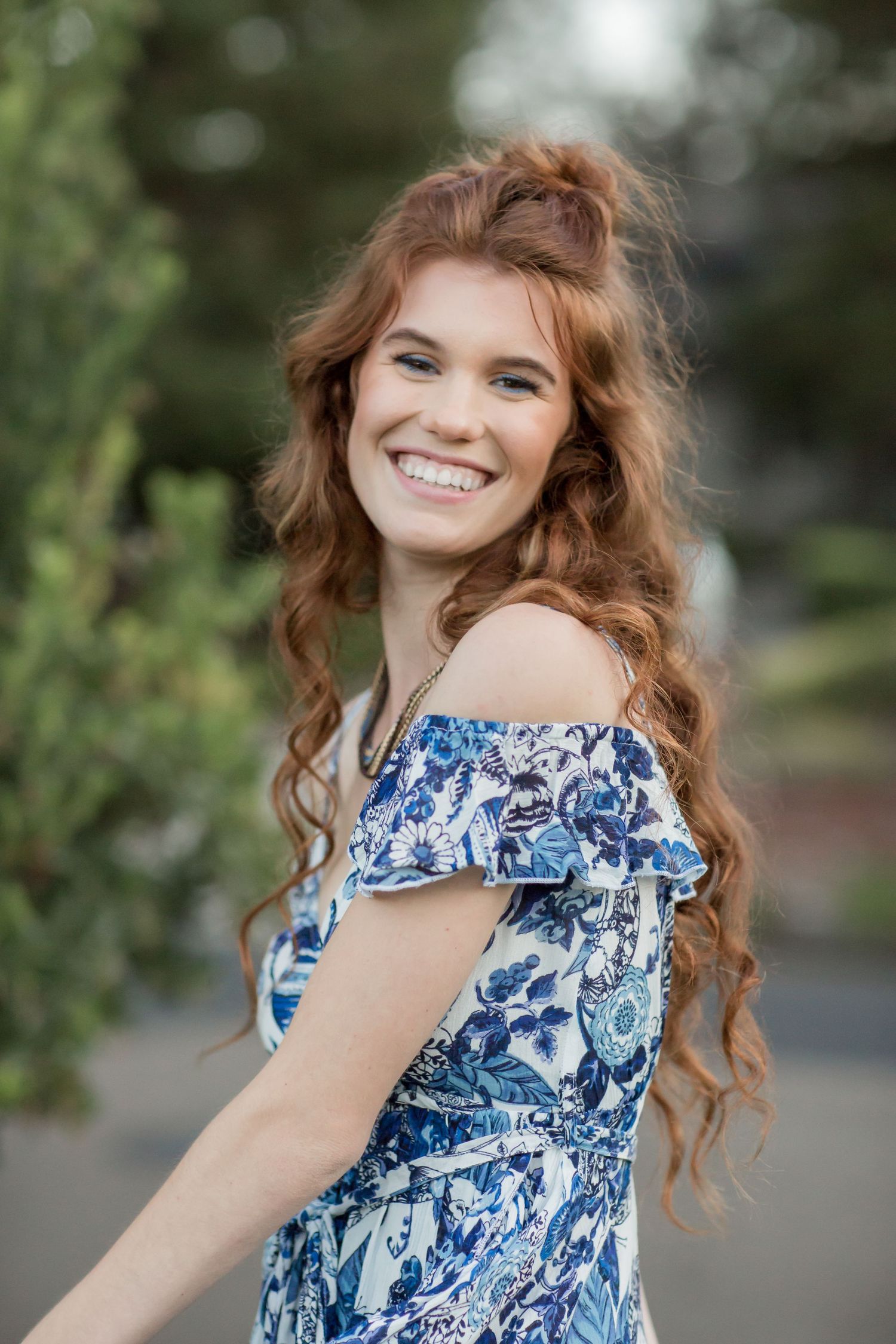 Smiling redhead young woman in blue floral off-shoulder dress with curly half-up hairstyle.