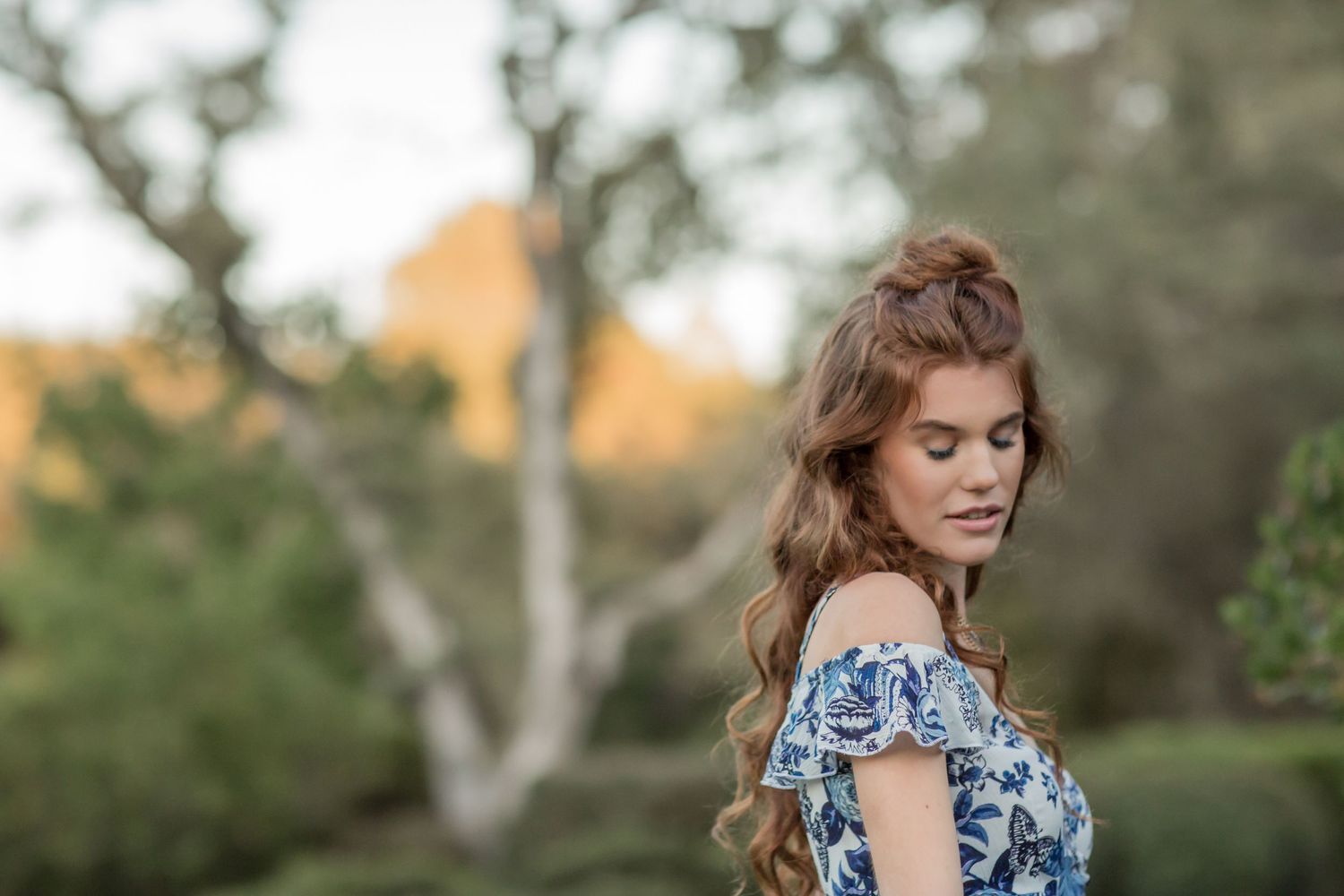 Young redhead woman in blue floral off-shoulder dress with elegant half-up hairstyle outdoors.