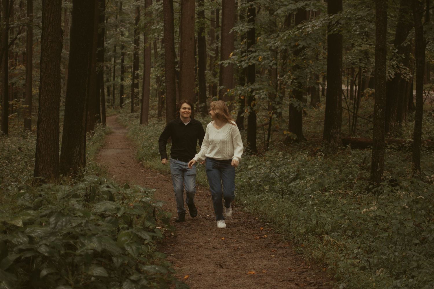 Couple walking hand in hand on a wooded trail through a lush forest.