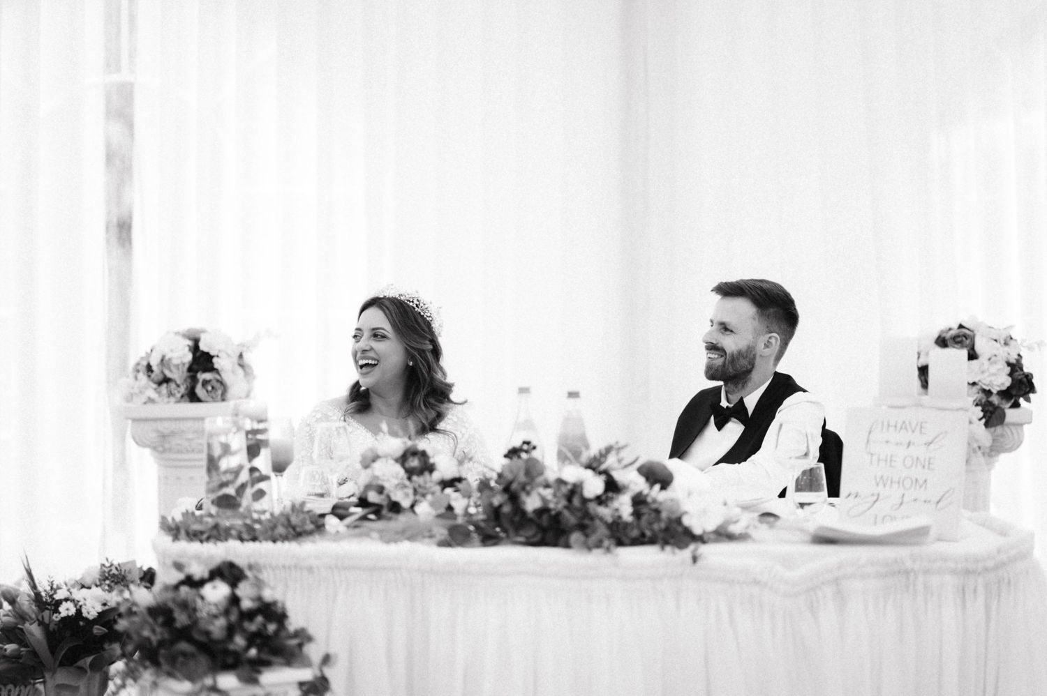 Black and white photo of a couple seated at their decorated wedding reception head table.