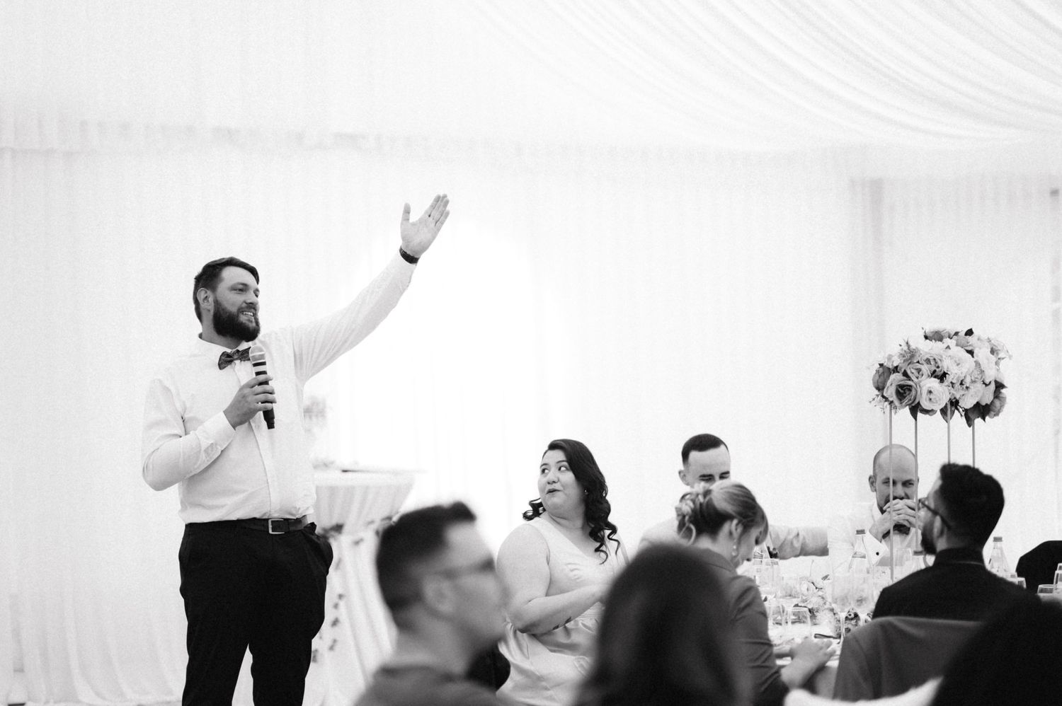 A black and white wedding reception scene with someone giving a speech at an indoor venue.