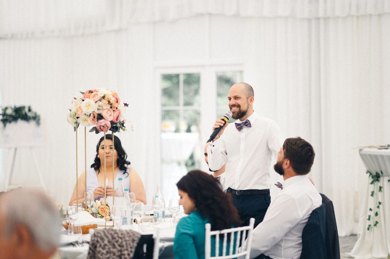 A wedding reception speech being given in a bright white indoor venue with floral decorations.
