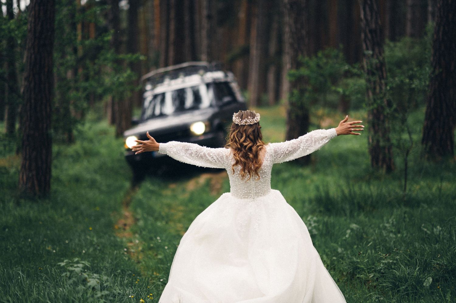 Bride in flowing white dress stands with outstretched arms in front of vintage vehicle in forest.