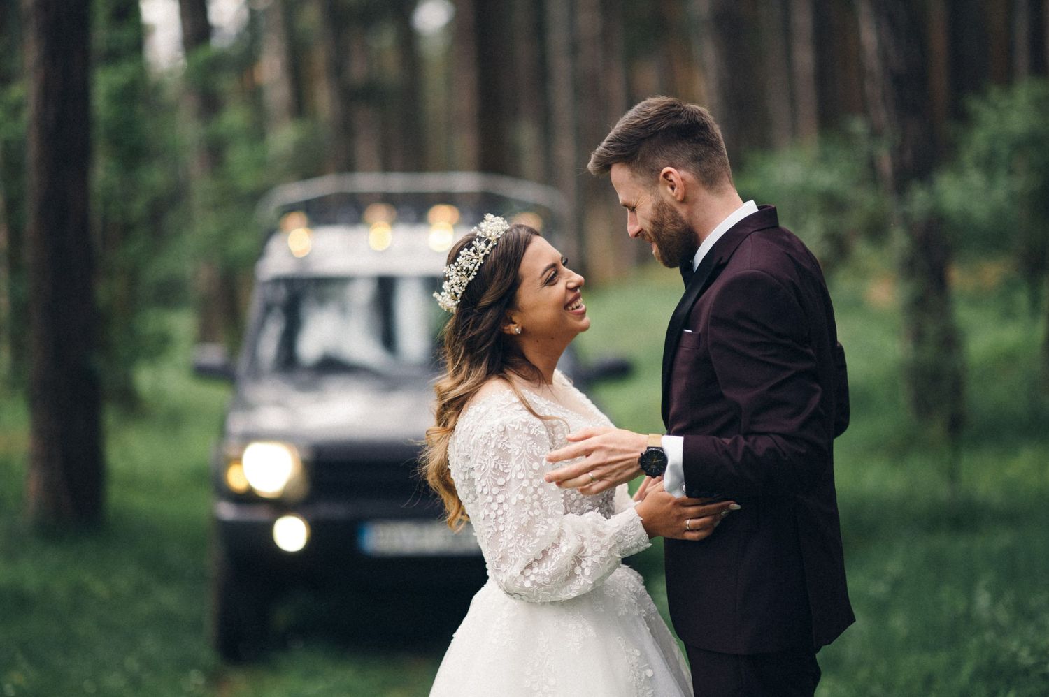 Wedding couple shares intimate moment in forest with classic Land Rover in background.