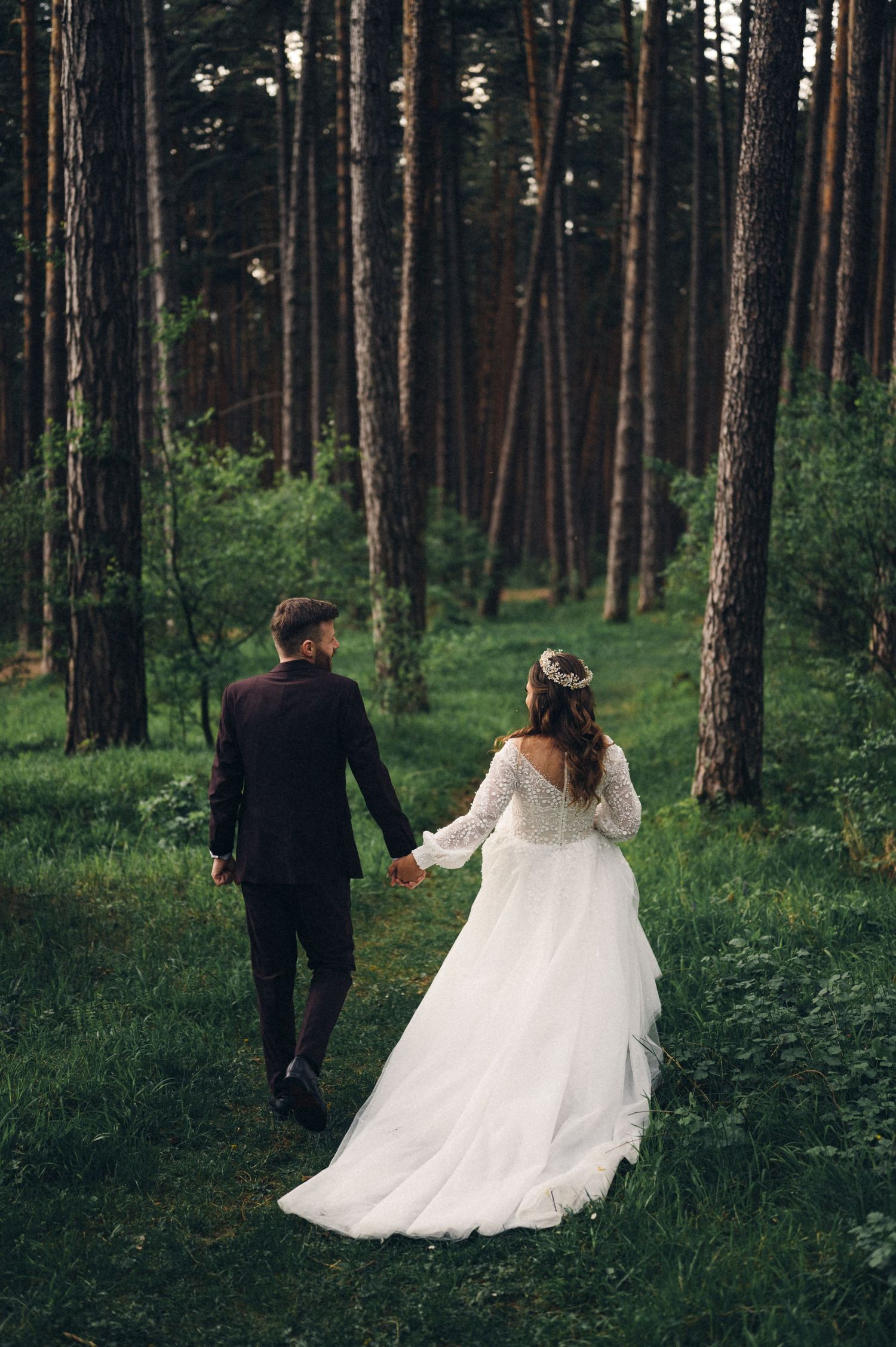 Newlyweds walking through pine forest holding hands on their wedding day.