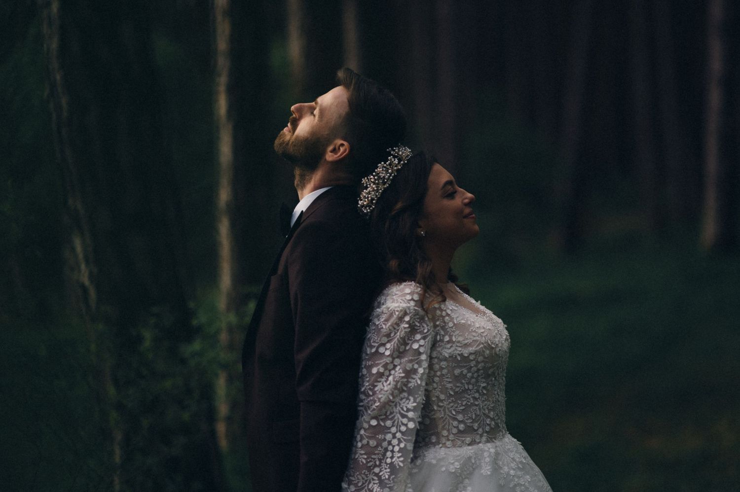 Bride and groom stand back-to-back in a dark forest, looking upward.