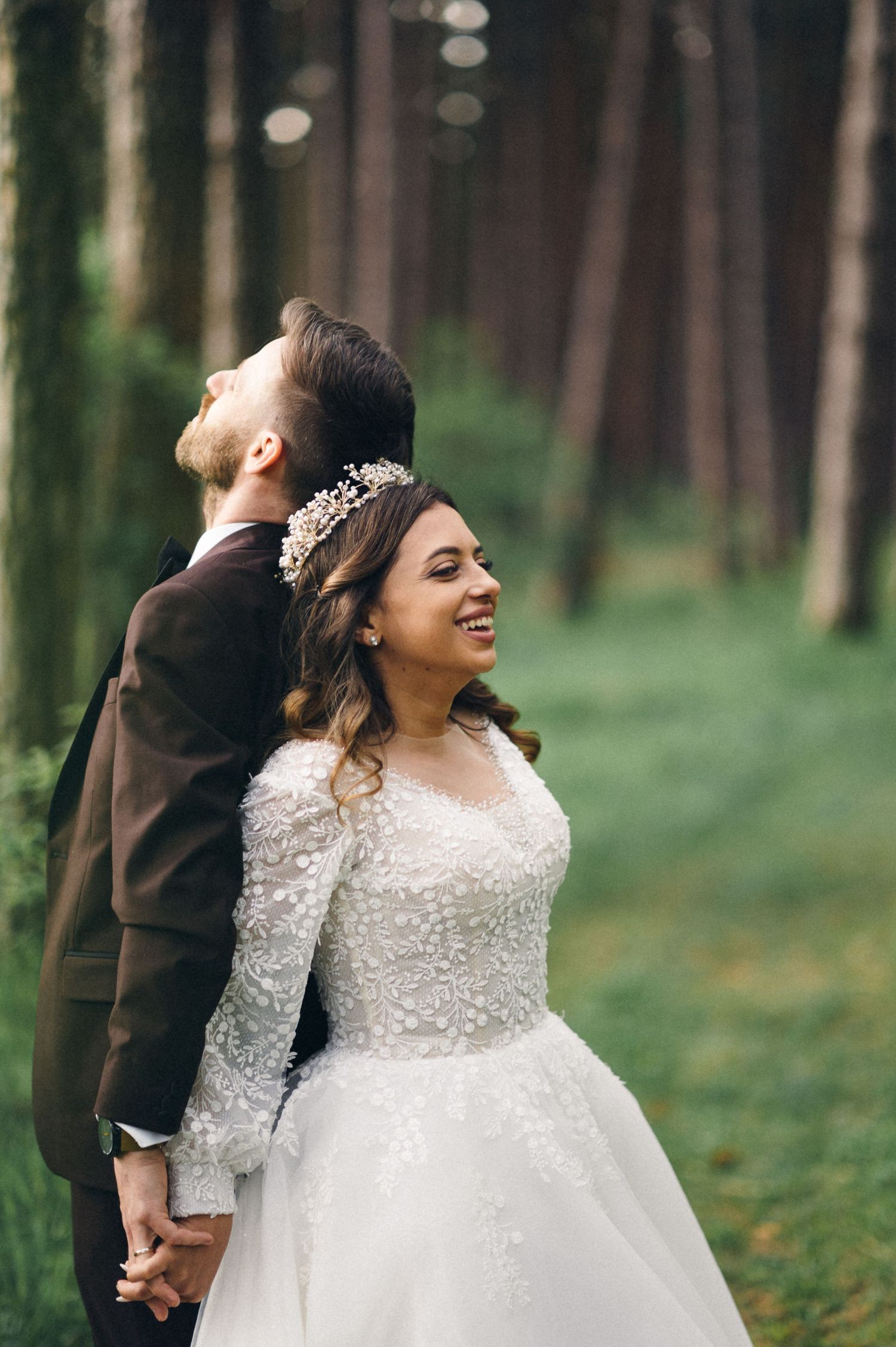 Bride in lace gown and groom in suit look up at forest canopy while holding hands.