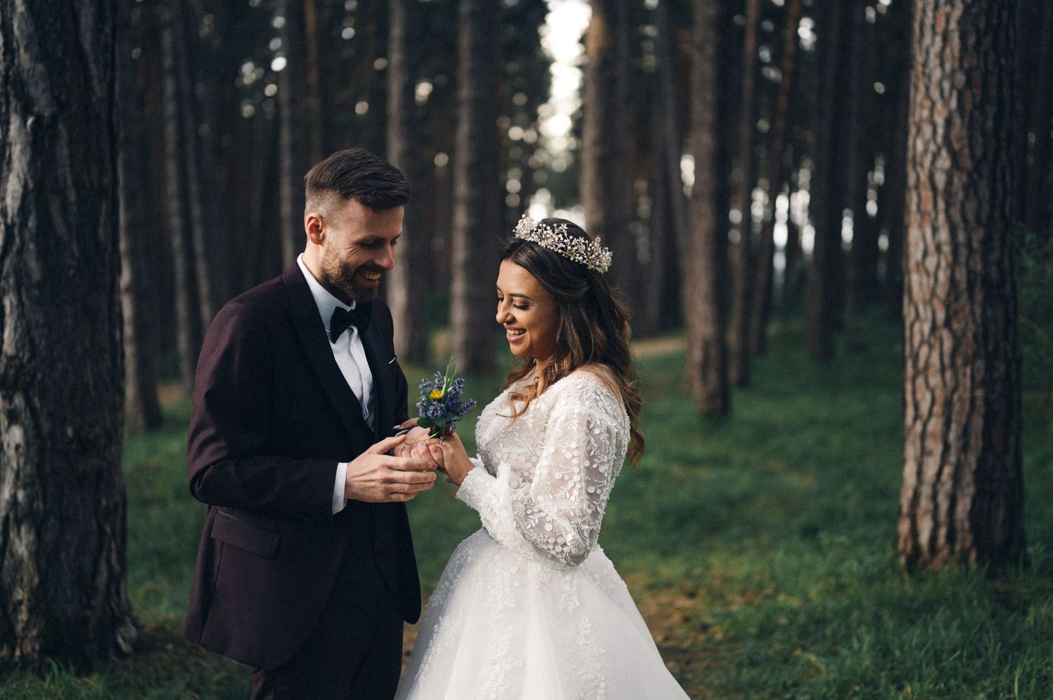 Bride in lace dress and groom in burgundy suit hold hands in a pine forest.