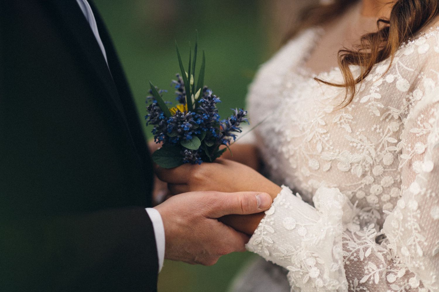 Close up detail of hands holding delicate blue wildflower bouquet against lace wedding dress.