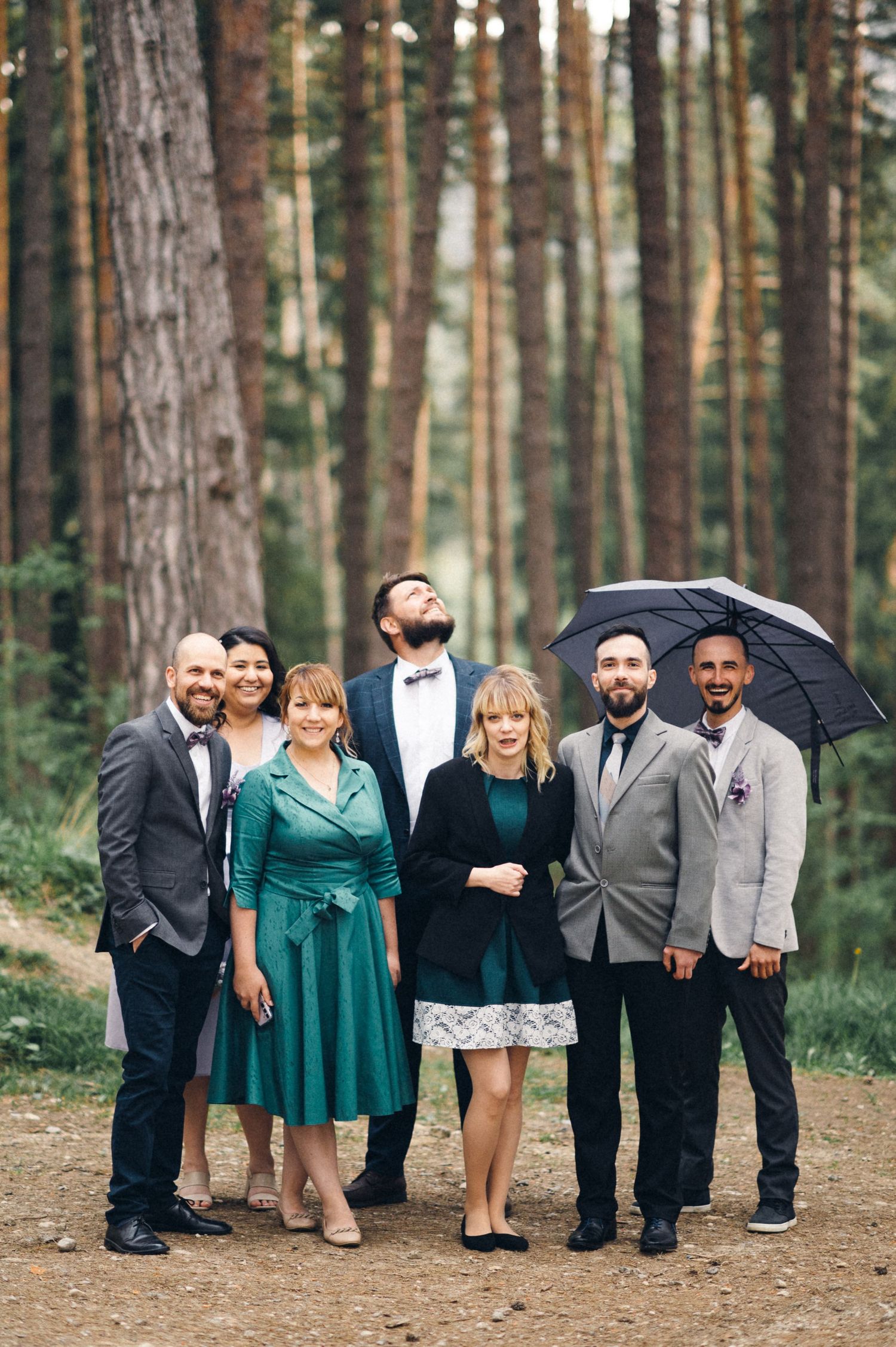 A wedding party poses together on a forest path with one person holding a black umbrella.