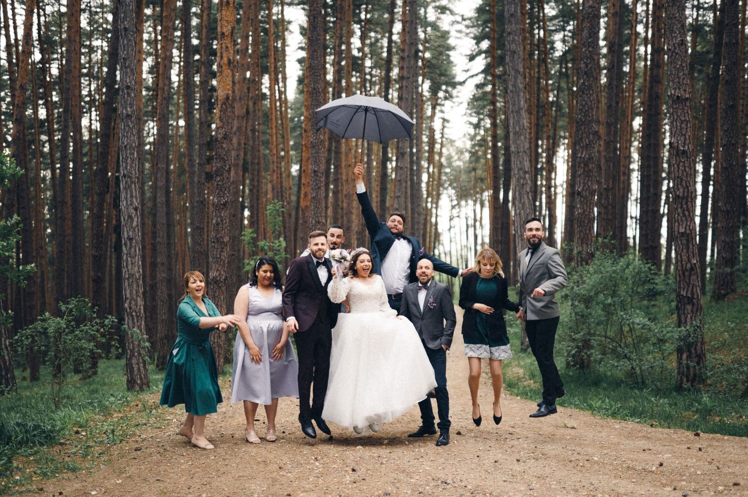 Wedding party members gather under an umbrella in a pine forest setting.