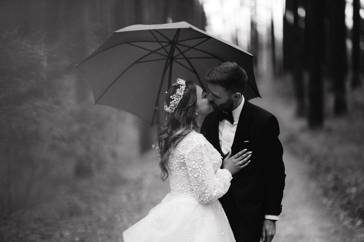 A black and white portrait of a couple sharing an intimate moment under an umbrella in the woods.