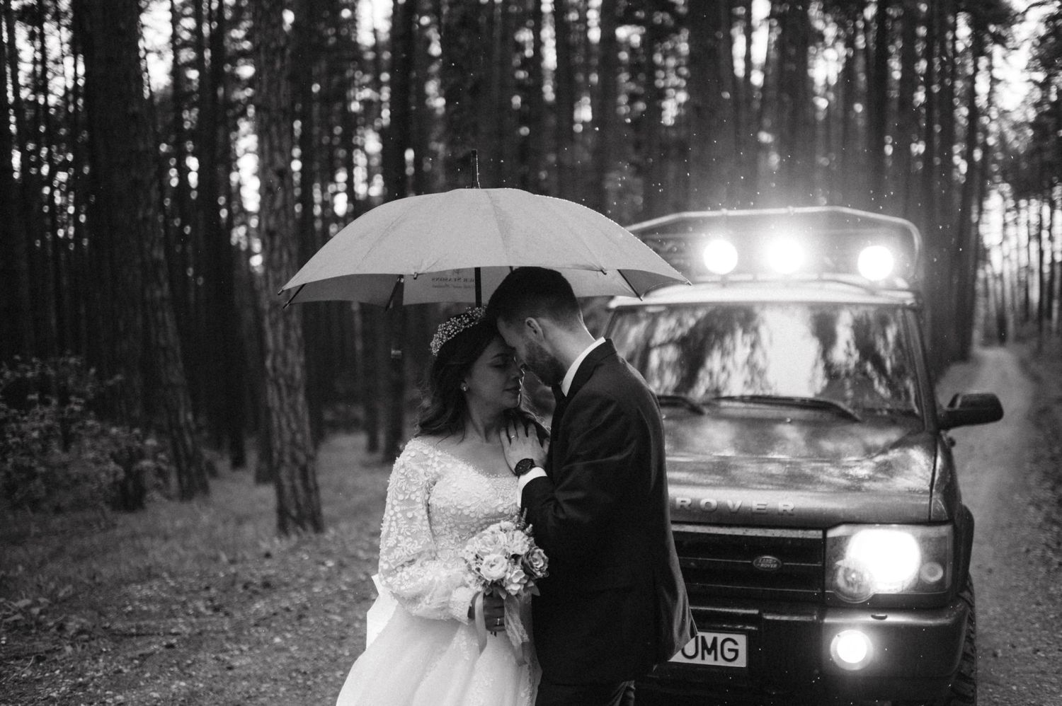 Romantic rainy wedding photoshoot with couple and vintage car in forest setting.