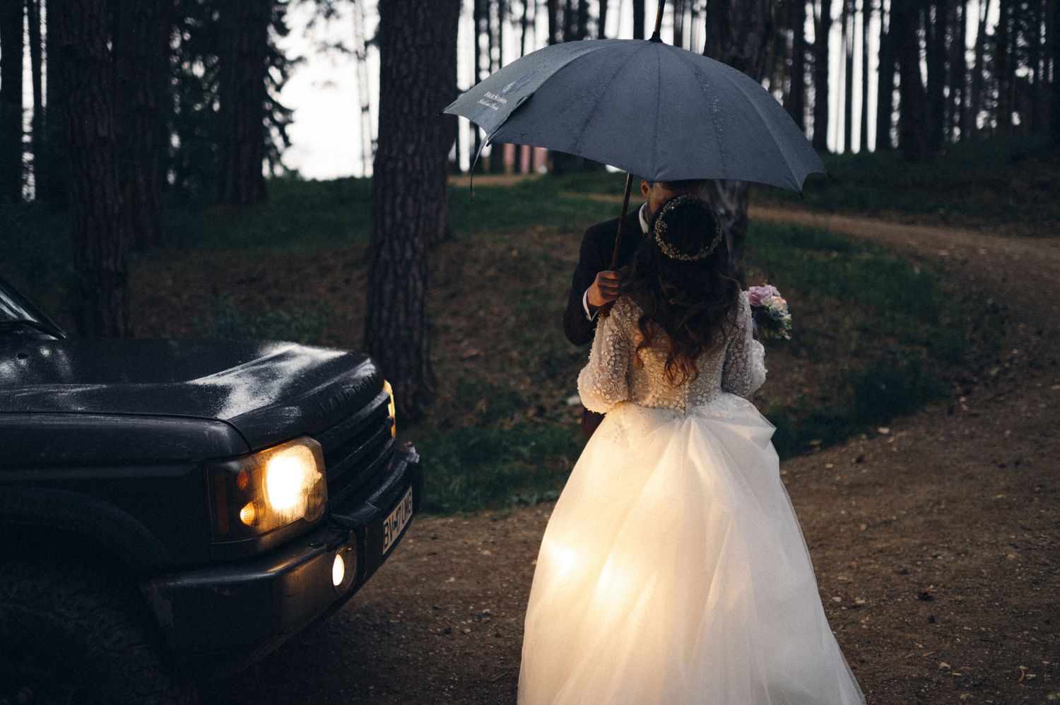 Silhouette of bride in flowing dress near car with umbrella at dusk.