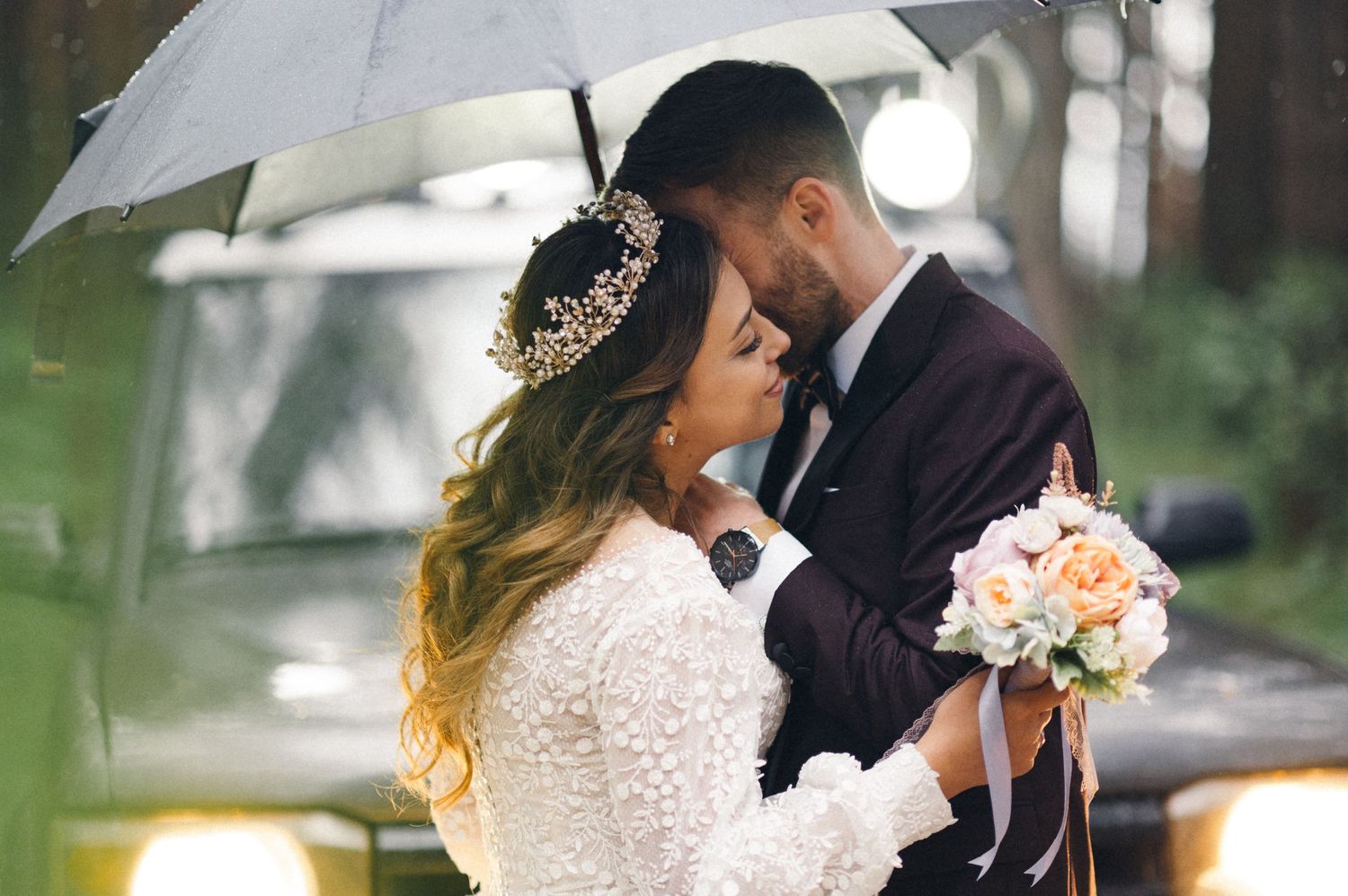 Tender embrace under white umbrella with bridal bouquet and lace dress details.